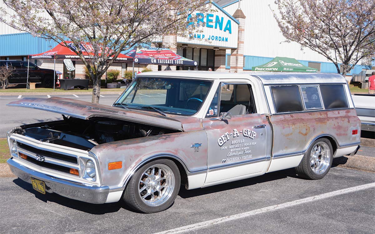 Landscape outdoor close-up photograph angle view of a vintage rusty faded light gray Chevrolet C10 truck vehicle parked in a parking lot area as there's a big building and other random assorted tents in the distant background