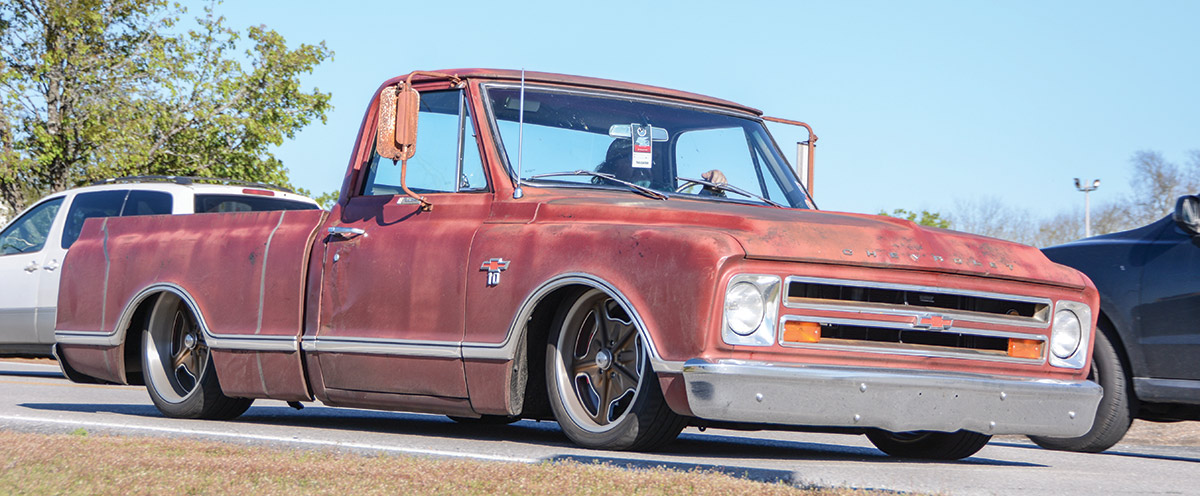 Landscape outdoor close-up photograph angle view of a vintage rusty faded dark red Chevrolet C10 truck vehicle with a guy driving the car on a road during the day
