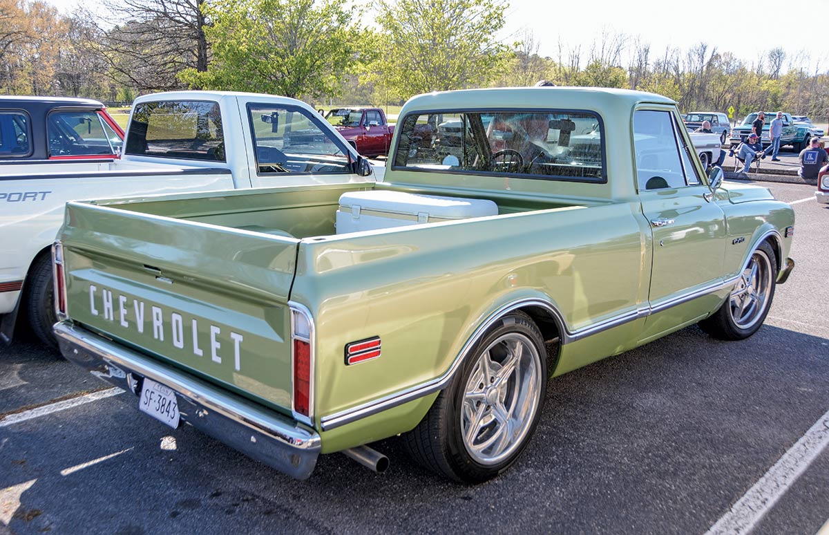 Landscape outdoor close-up photograph rear angle view of a vintage faded light metallic lime green colored Chevrolet truck vehicle parked in a parking lot area nearby other random assorted cars during the day