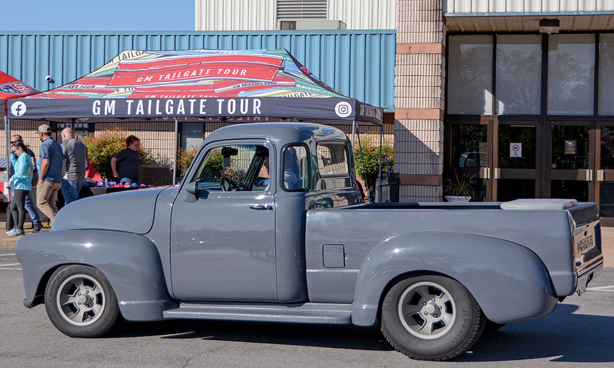Landscape outdoor close-up photograph side view of a vintage metallic dark grey Chevrolet truck vehicle parked nearby a building, a tent, and other random people around as there is a man in the driver's seat of the car