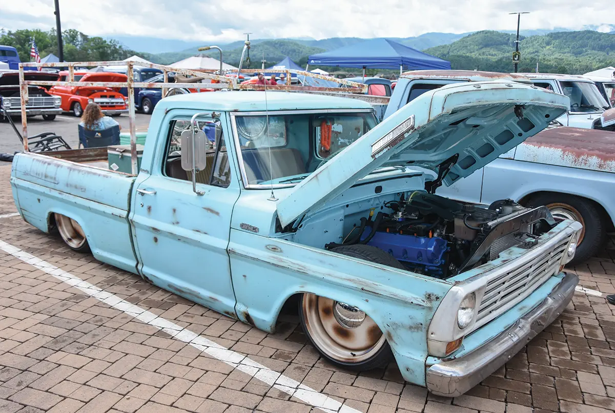 Landscape close-up photograph view of a vintage rusty light powder blue Ford F-100 truck vehicle with its front top hood slightly open halfways revealing its exposed engine as it is parked on a brick ground parking lot area nearby other random assorted vehicles and tents in the far distance on a somewhat cloudy overcast day