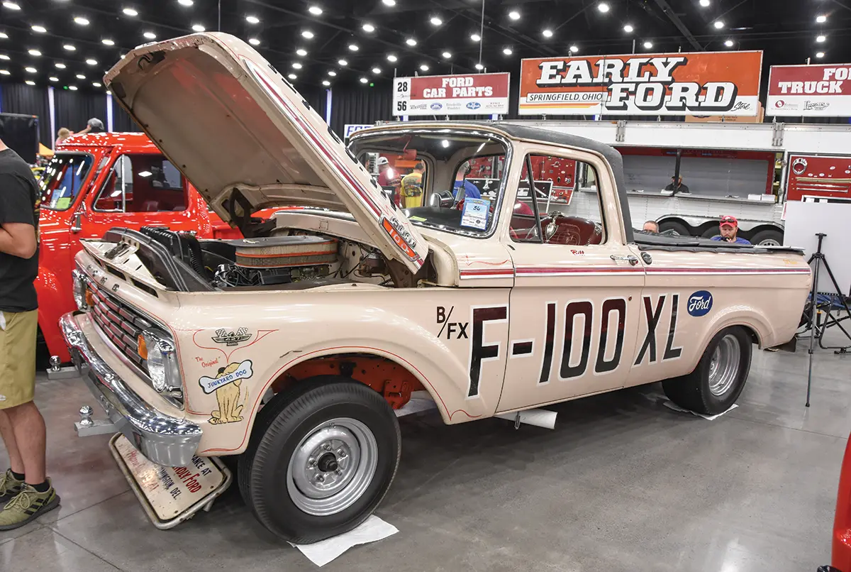 Landscape close-up photograph view of a custom-themed vintage metallic dark beige/red/white/black Ford F-100 truck vehicle with its front top hood slightly open halfways revealing its exposed engine plus across the driver's door area it shows B/FX F-100 XL as well as the Ford logo and on the side of the grille area it shows The Original Junkyard Dog painting illustration as this car is parked inside a showroom area of a building as there are people nearby and other signage banners in the far distance