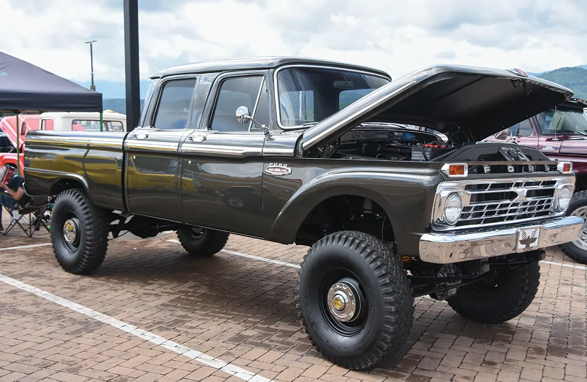 Landscape close-up photograph view of a vintage metallic black Ford F-250 four wheel drive truck vehicle with its front top hood slightly open halfways revealing its exposed engine as it is parked on a brick ground parking lot area nearby other random assorted vehicles, a person seated down in a tent chair, and a tent in the far distance on a somewhat cloudy overcast day