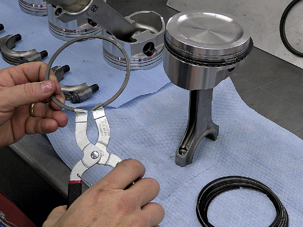 Piston ring installation in progress on a forged piston, with connecting rods and clips nearby
