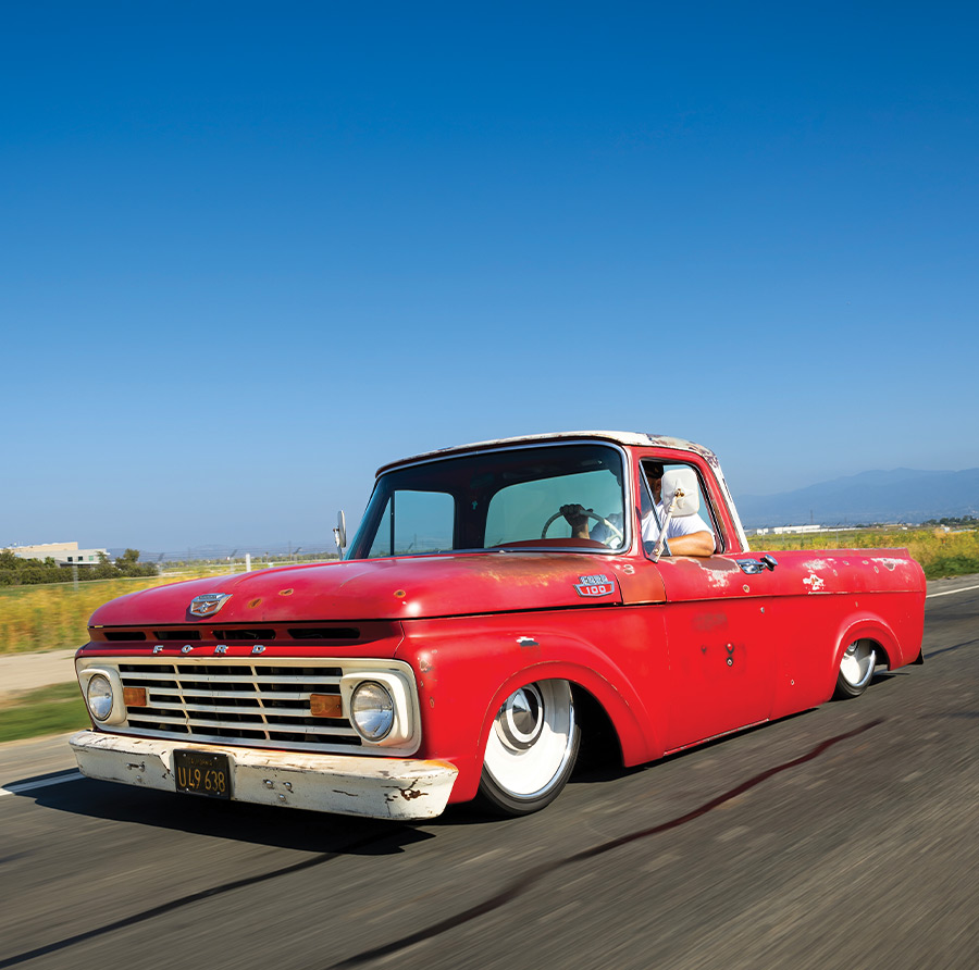 A vibrant red Ford F-100 cruising on a scenic open road, slammed to the ground on white steel wheels