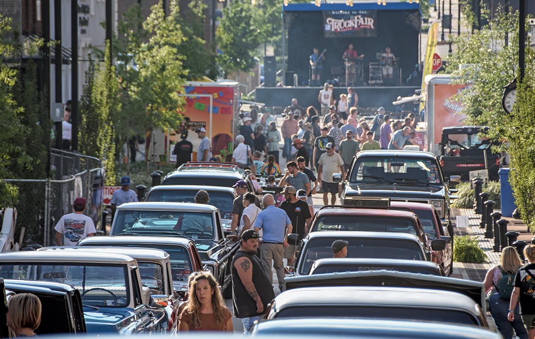 crowded truck show street scene with a stage and classic trucks lined up