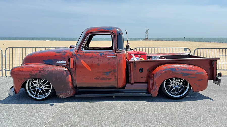 Side profile of a vintage Chevrolet truck with a distressed, rusted paint job and a custom chassis, on display at a seaside car show.
