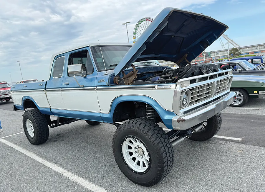 Lifted two-tone blue and white Ford pickup truck with large off-road tires and its hood open, on display at an outdoor show.