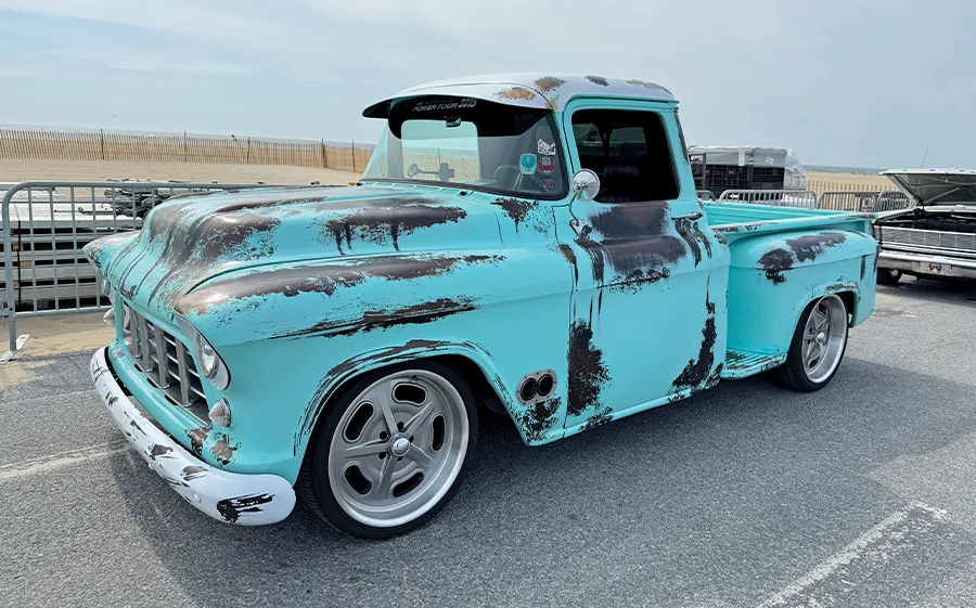 Vintage Chevrolet truck with a custom, faded blue-green paint job and rusty accents, on display at an outdoor car show with the ocean in the background.