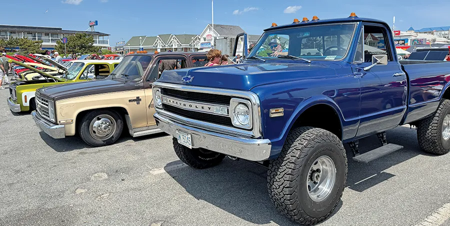 Lifted blue classic Chevrolet pickup truck with large off-road tires, parked alongside other vintage trucks at a sunny car show.