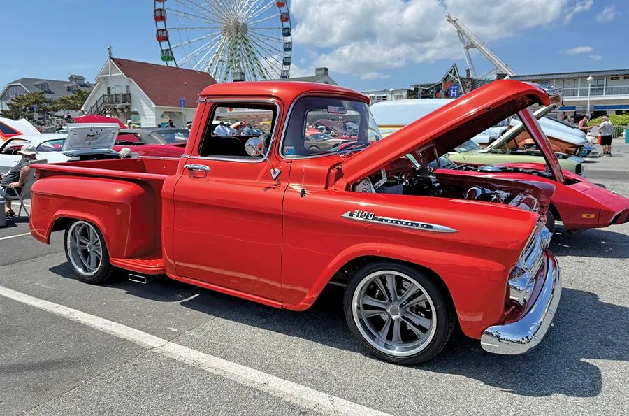 Vintage Chevrolet pickup truck with a glossy red paint job, on display with its engine bay exposed at a sunny seaside car show.