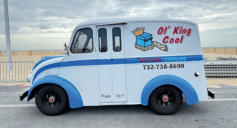 Restored vintage white and blue heating and cooling service van 'Ol' King Cool' with a lowered stance, parked on a street near the ocean.