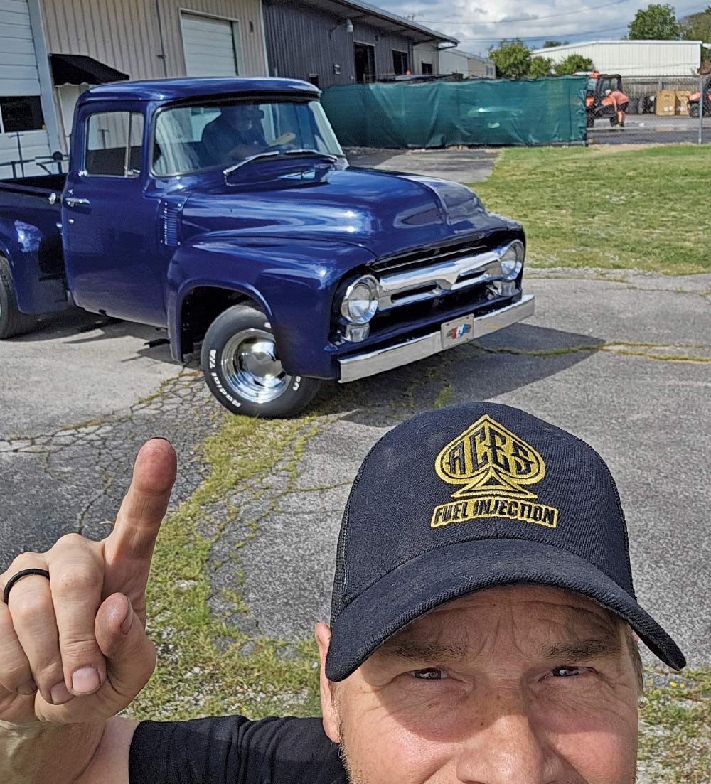 A low-angle selfie of a man pointing at a blue, classic Ford F-100 pickup truck parked behind him.