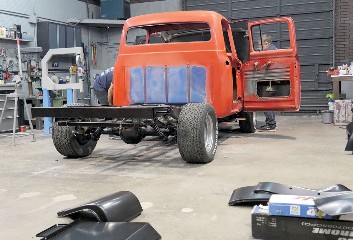 A wide-angle interior shot of a workshop, showing the rear of a red classic Ford pickup truck in the midst of restoration.