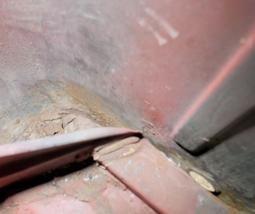 A close-up, low-angle shot of a rusted and damaged section of a red vehicle's metal paneling. 