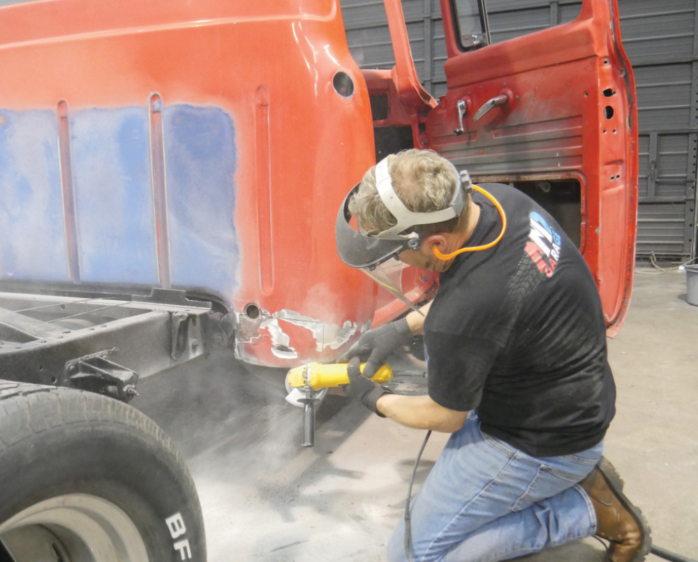 A person wearing a face shield and ear protection kneels on the floor, using a grinder to work on the metal body of a red classic Ford pickup truck.