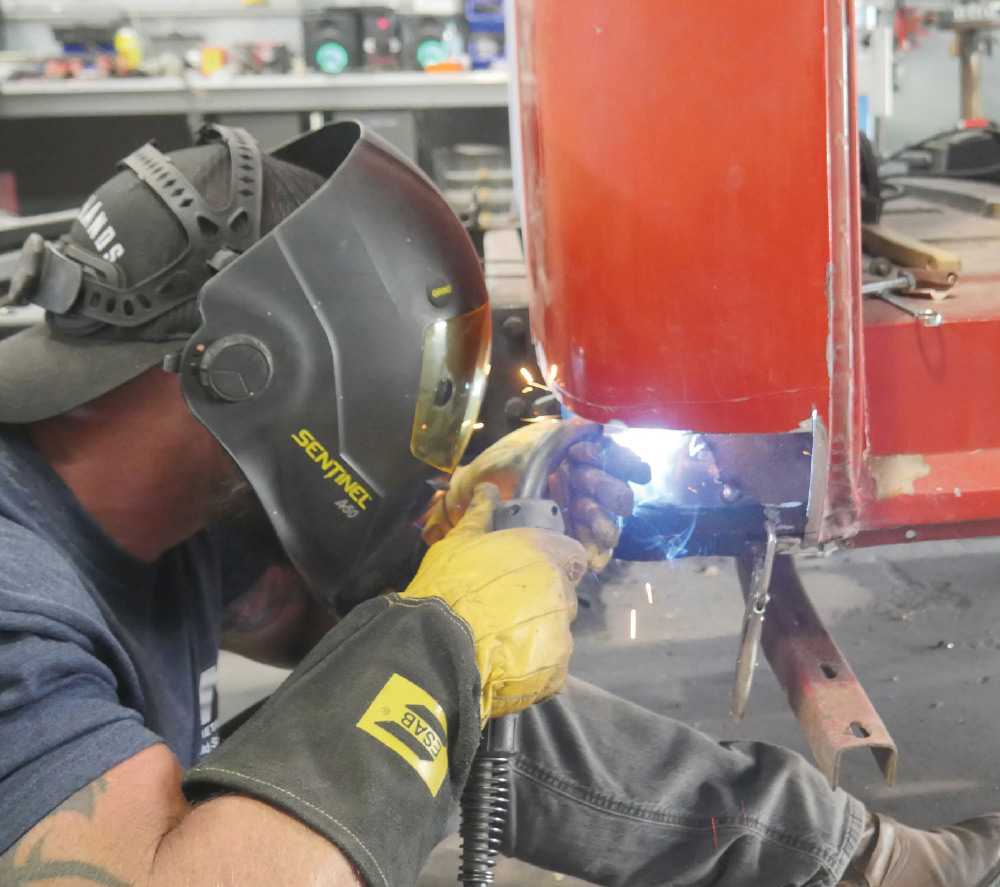 A man wearing a welding helmet and protective gloves is welding a new metal patch panel onto a red classic pickup truck.