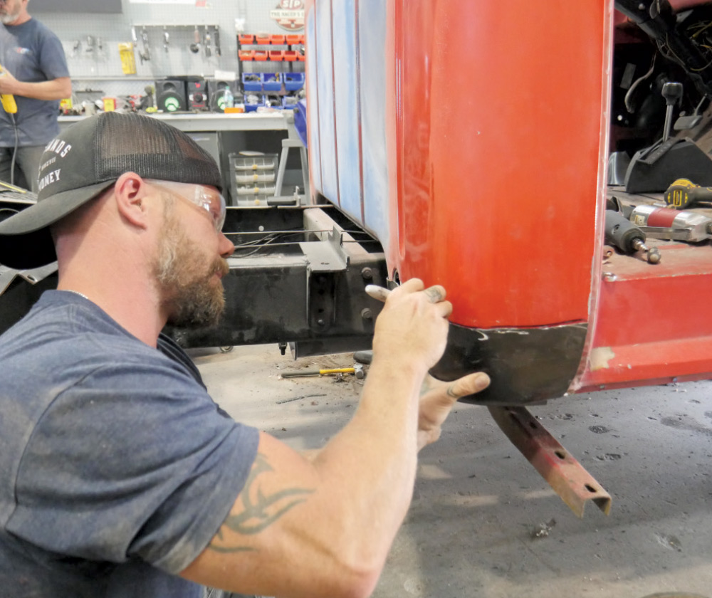A person wearing a backward-facing cap and safety glasses kneels on the floor, using a tool to work on the newly installed patch panel on a red classic pickup truck. 