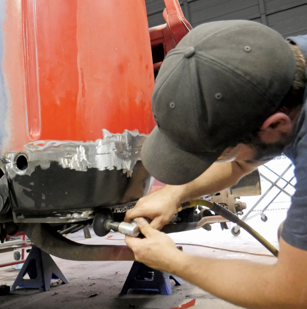 A medium shot of a person wearing a backward-facing cap, using a tool to smooth out a newly welded patch panel on a red classic pickup truck. 