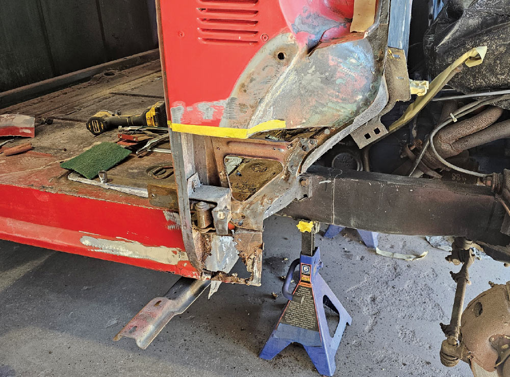 A low-angle close-up shot of the front-left section of a red classic pickup truck's cab, showing extensive disassembly and rust damage.