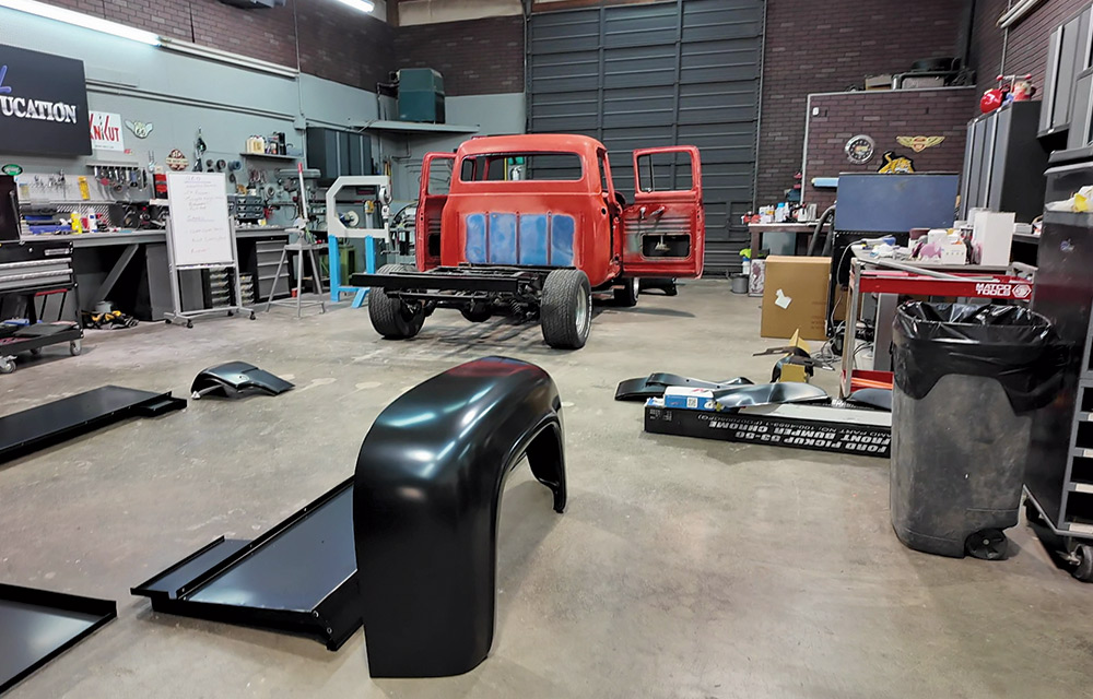 A wide-angle interior shot of a workshop, with the rear of a red classic pickup truck under restoration in the center. 