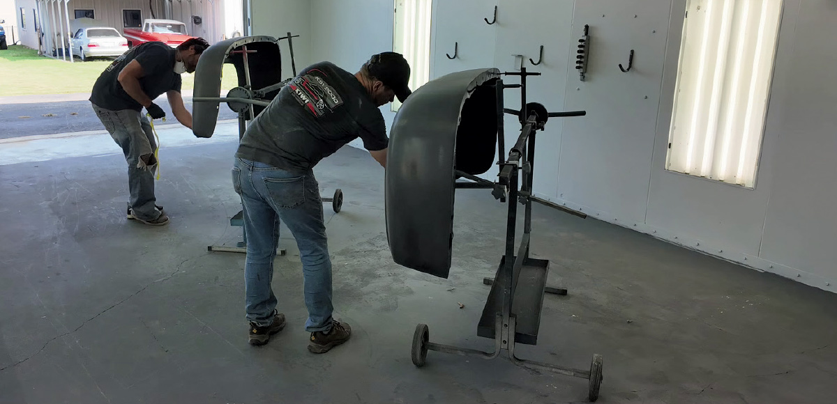 A wide-angle interior shot of a workshop or paint booth, showing two men working on classic truck fenders. 