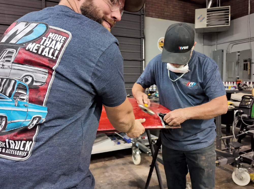 A close-up interior shot of two men in a workshop, working together on a red metal panel.