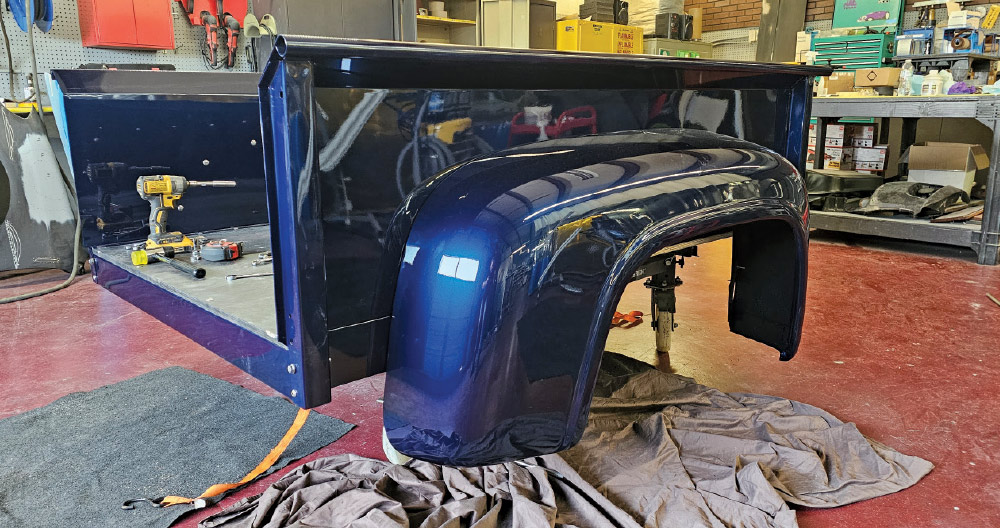 A low-angle interior shot of a workshop, showcasing a newly painted dark blue classic truck bed and fender.
