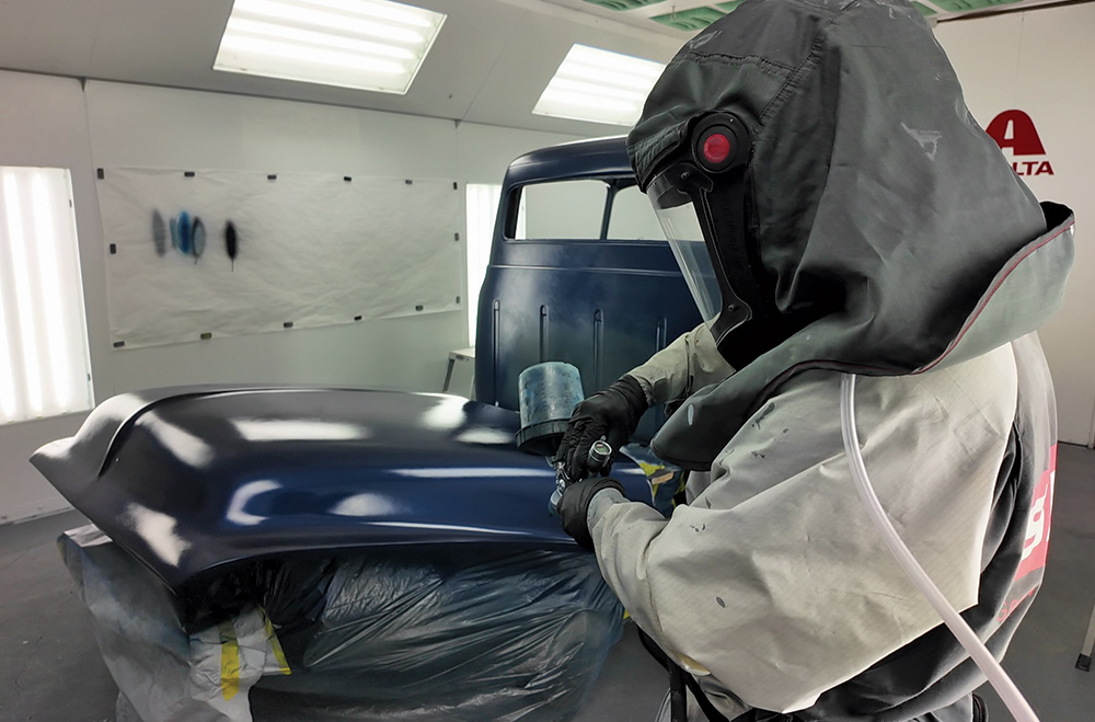 A medium interior shot of a paint booth, showing a person in a full protective suit and respirator using a spray gun to apply a glossy, dark blue coat of paint to a classic truck's hood.