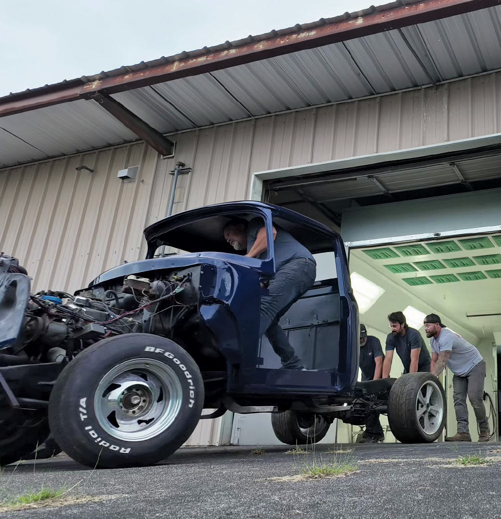A wide-angle, low-angle shot of three men pushing a dark blue classic pickup truck's cab and chassis out of a garage.
