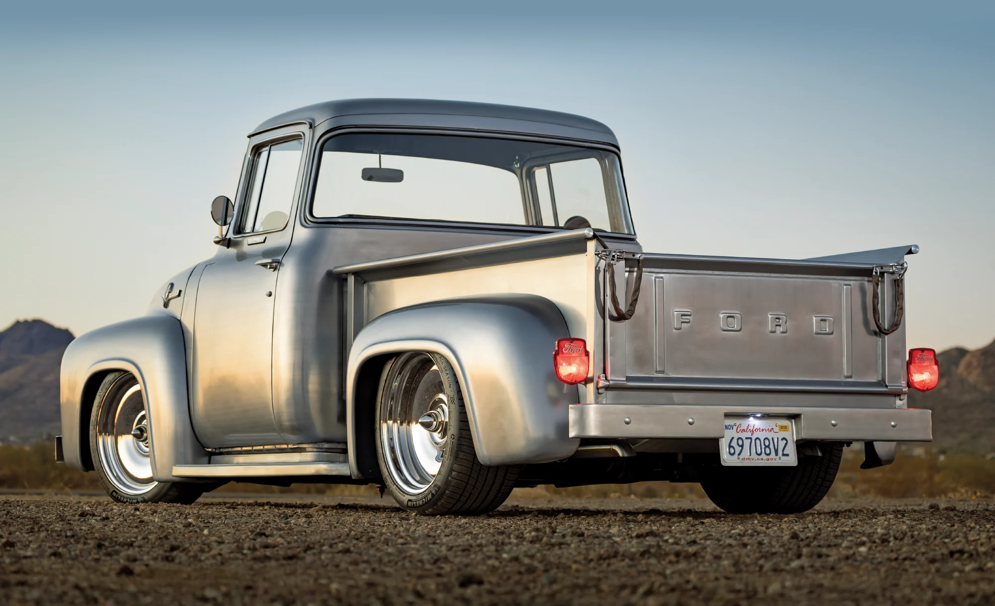 A three-quarter rear view of a custom 1956 Ford F-100 classic pickup truck parked on a dirt road in a desert landscape at sunset. 