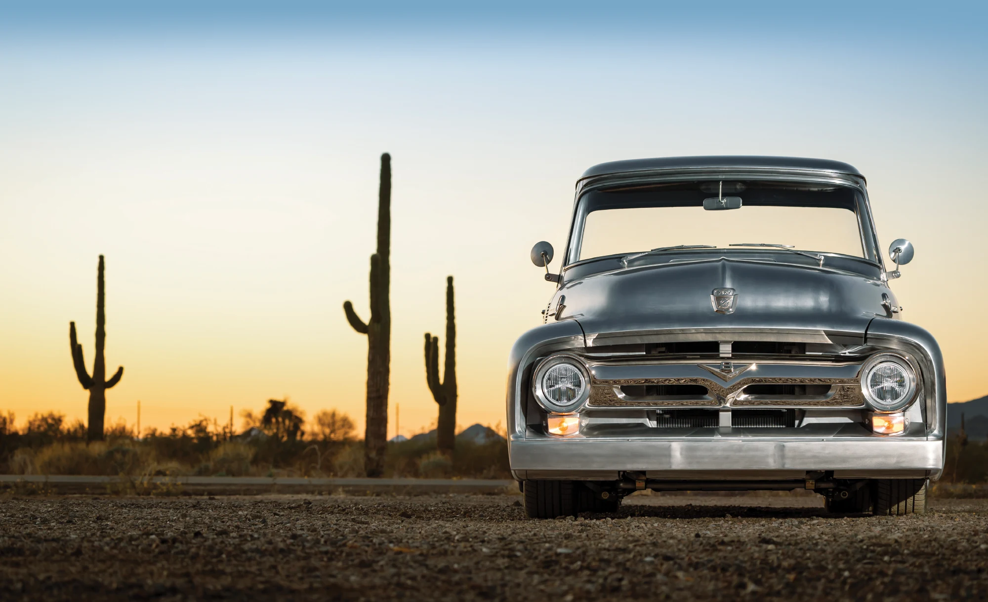 A straight-on front view of a custom 1956 Ford F-100 classic pickup truck parked on a dirt road in a desert landscape at sunset. 