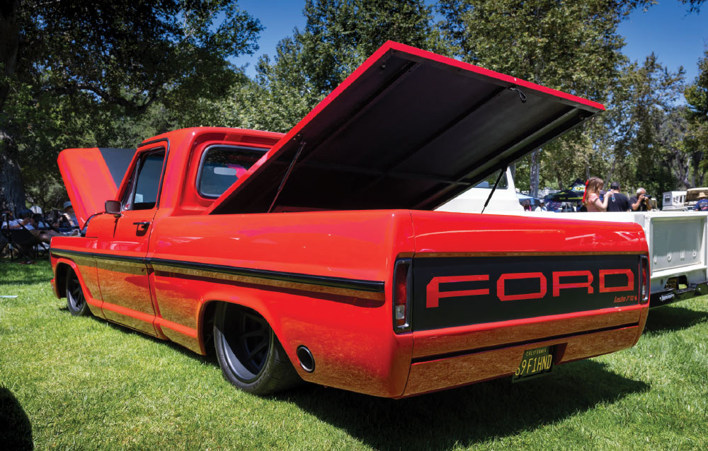 A low-angle, rear-side view of a classic red Ford pickup truck, likely an F-Series, that has been highly customized. It is lowered close to the ground, features black custom wheels, and has the tailgate removed and replaced with a smoothed rear panel that has black "FORD" lettering. The bed cover is hinged and propped open. The truck is parked on a sunny, grassy field.