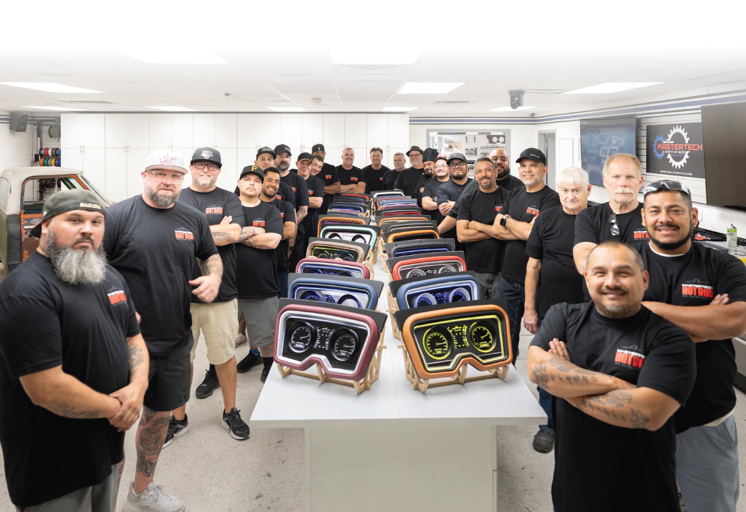 A large group of men, many wearing black "HOT ROD" t-shirts, stands in a brightly lit workshop. They are gathered behind a table displaying several colorful, custom-built vehicle dashboards. The men are smiling and looking at the camera. A couple of classic car bodies are visible in the background on the left.