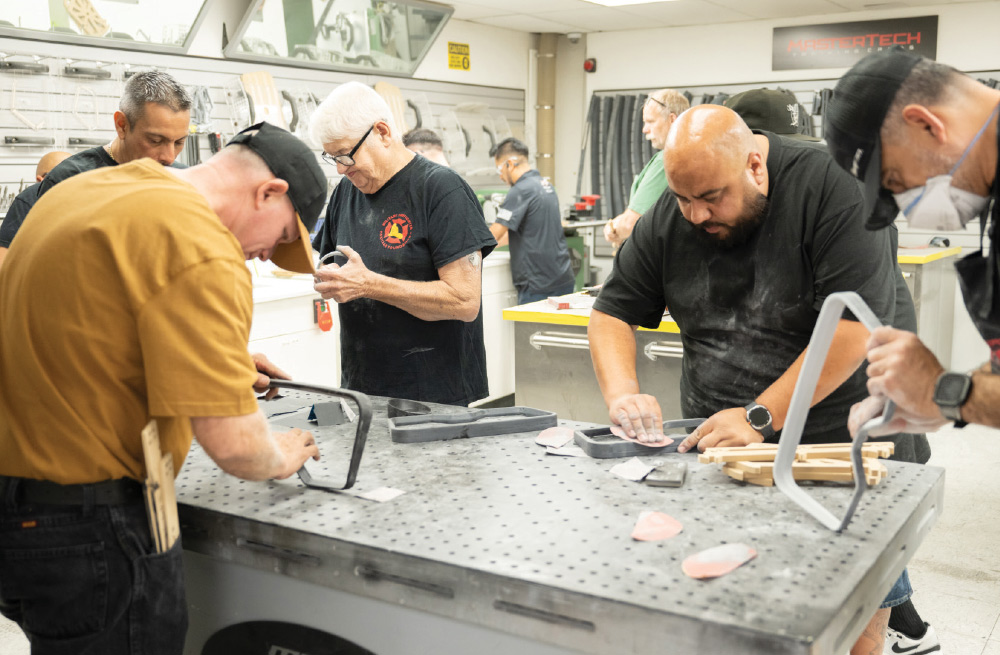 A group of men in a workshop is gathered around a large metal workbench, focusing on tasks involving sanding and preparing parts. The man in the foreground on the right is sanding a dark, curved component. Other men are similarly engaged in working on pieces of custom automotive parts.