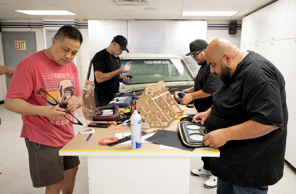 Four men are working on automotive projects in a brightly lit workshop. In the foreground, a man in a red t-shirt examines a pair of glasses while three other men, wearing black shirts, are gathered around a table assembling custom dashboard components. A white vintage pickup truck is visible in the background.