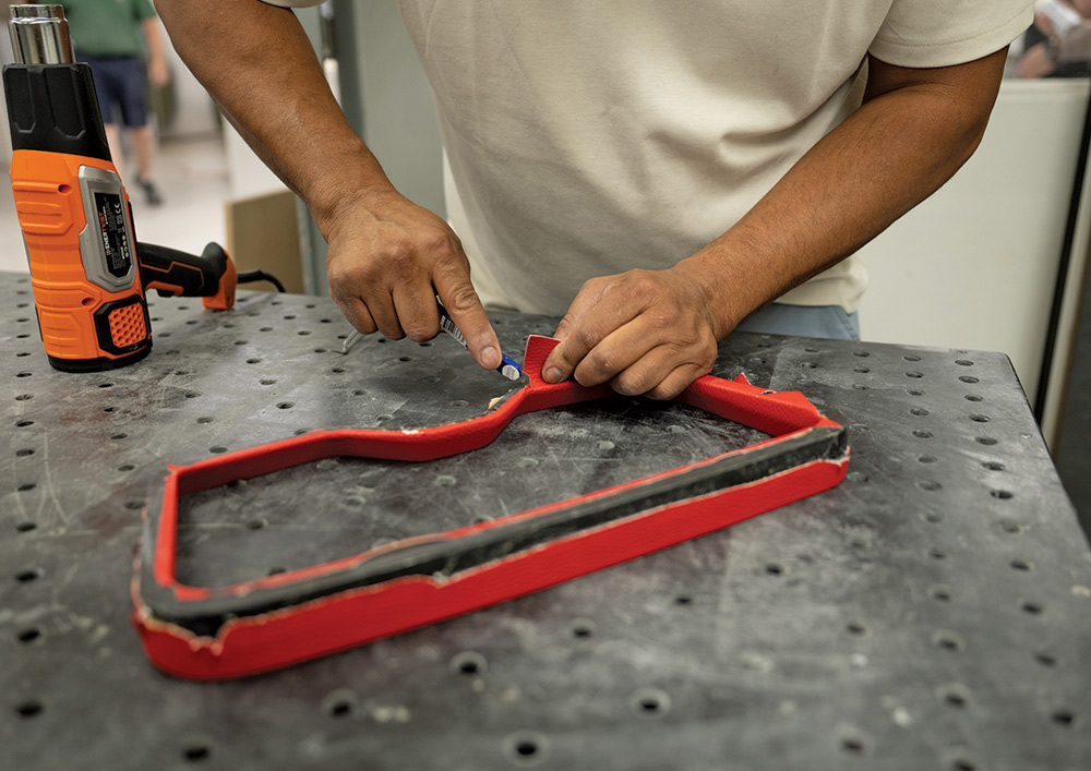 A close-up shot of a person's hands working on a custom car part. The person, wearing a light-colored shirt, is using a small tool to carefully trim or tuck the edge of a piece of red vinyl or leather upholstery wrapped around a black, rectangular bezel. An orange and black heat gun is visible on the work surface to the left.