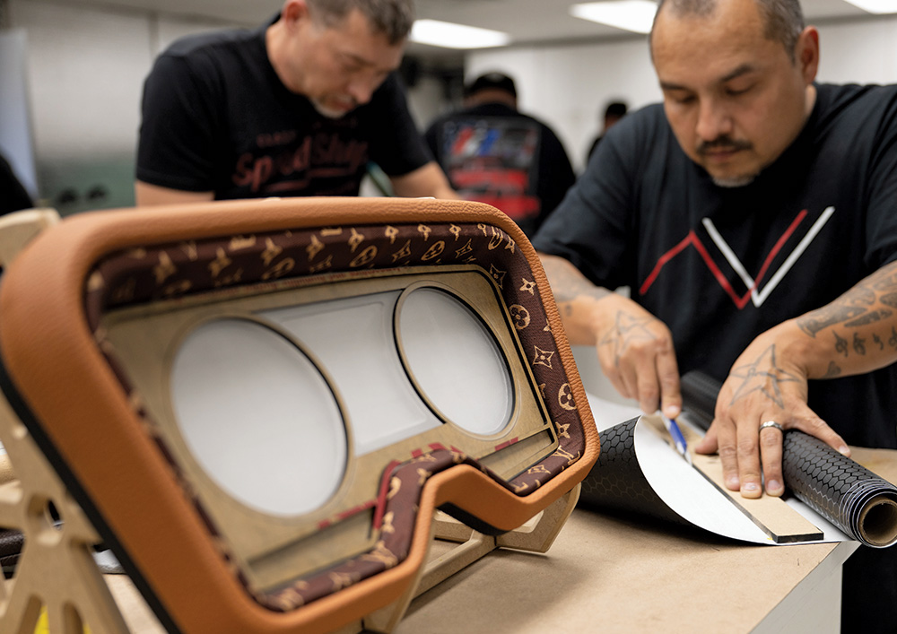 A close-up of a custom vehicle dashboard being upholstered. The dashboard frame, which has two large circular openings for gauges, is wrapped in tan material and an inner bezel features a patterned fabric. In the foreground, a man is focused on cutting a piece of black patterned vinyl from a roll, while another man works in the background.