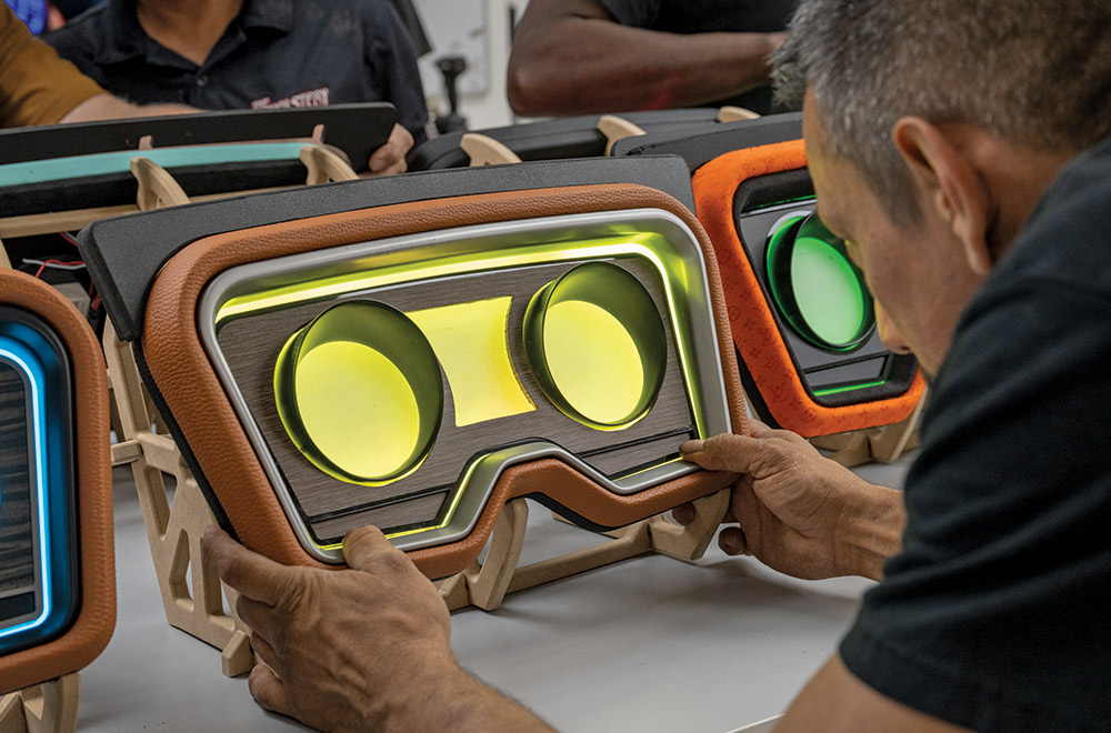 A close-up of a man examining a custom vehicle dashboard unit that is illuminated with bright yellow-green light. The dashboard is upholstered in tan leather with a black top and features a simulated woodgrain center panel with two circular gauge openings. Several other colorful custom dashboards are visible lined up on the table next to it.
