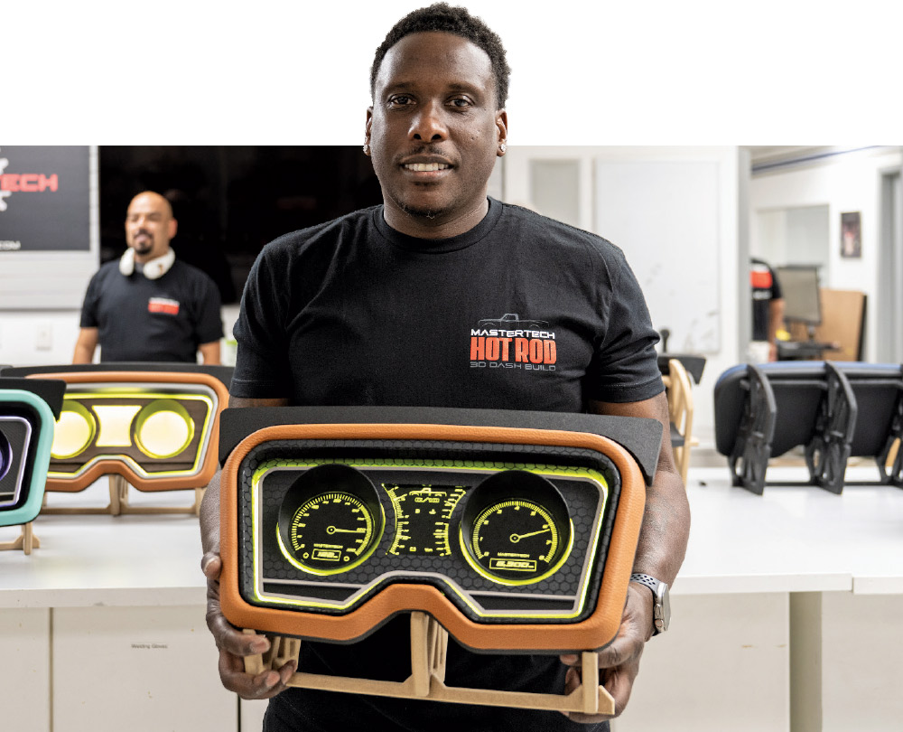A man wearing a black t-shirt with "MASTERTECH HOT ROD DASH BUILD" printed on it holds up a custom-built vehicle dashboard. The dashboard features a tan upholstered frame, a dark center panel, and two bright yellow-green digital gauges. He is smiling and looking directly at the camera. Another man is visible in the background.