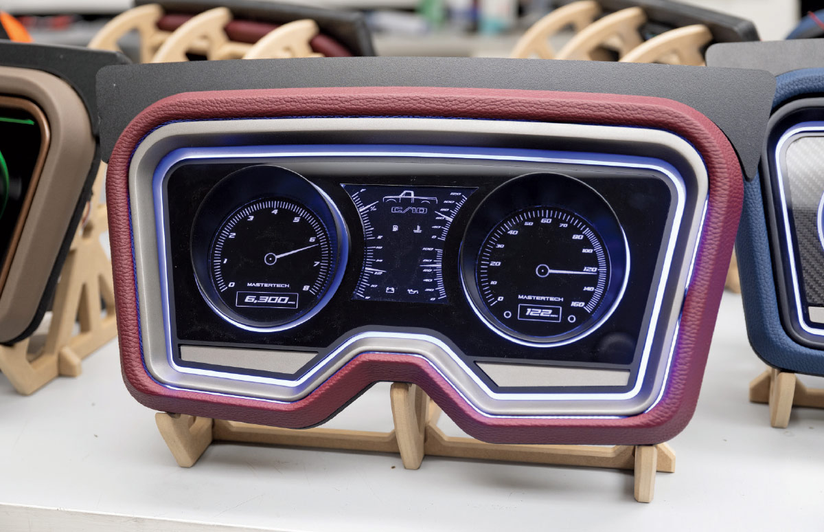 A close-up of a custom vehicle dashboard unit. The frame is upholstered in a dark red textured material, and the inner bezel is silver with a white/light blue neon glow border. The center panel is black and features two digital gauges with white needles and markings and a screen in the middle, all glowing with a cool white light. It rests on a wooden display stand.