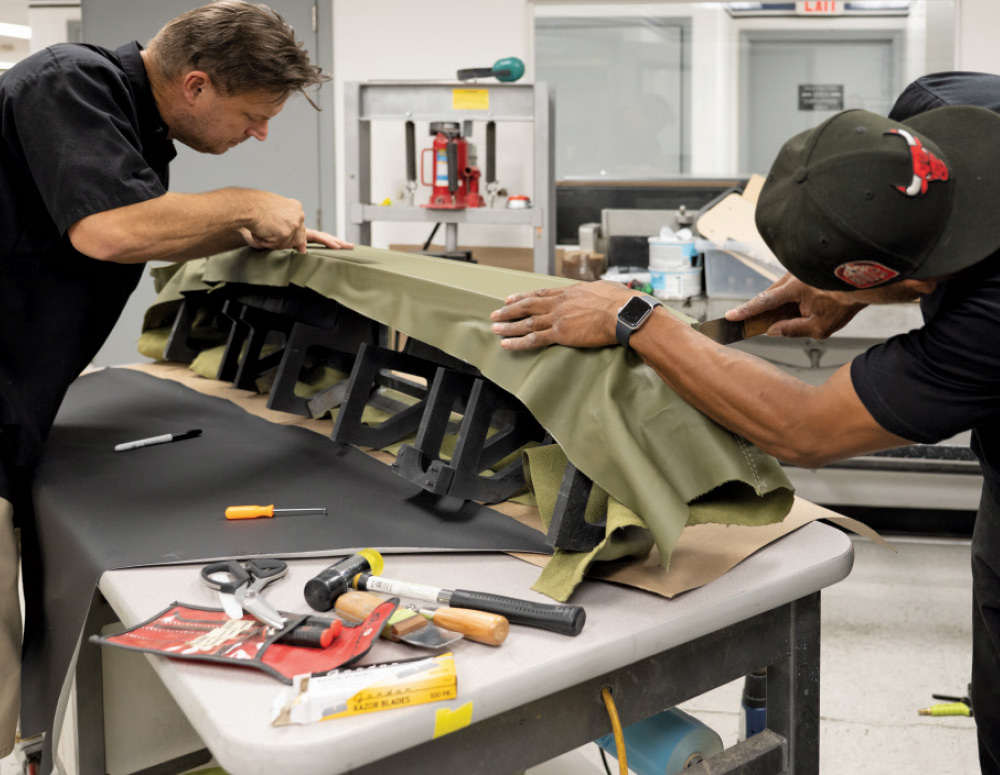 Two men are working together to upholster a large, custom vehicle dash component with olive green fabric or vinyl. They are standing over a workbench that holds the component on a black frame. Various tools, including scissors, a hammer, and razor blades, are scattered in the foreground.