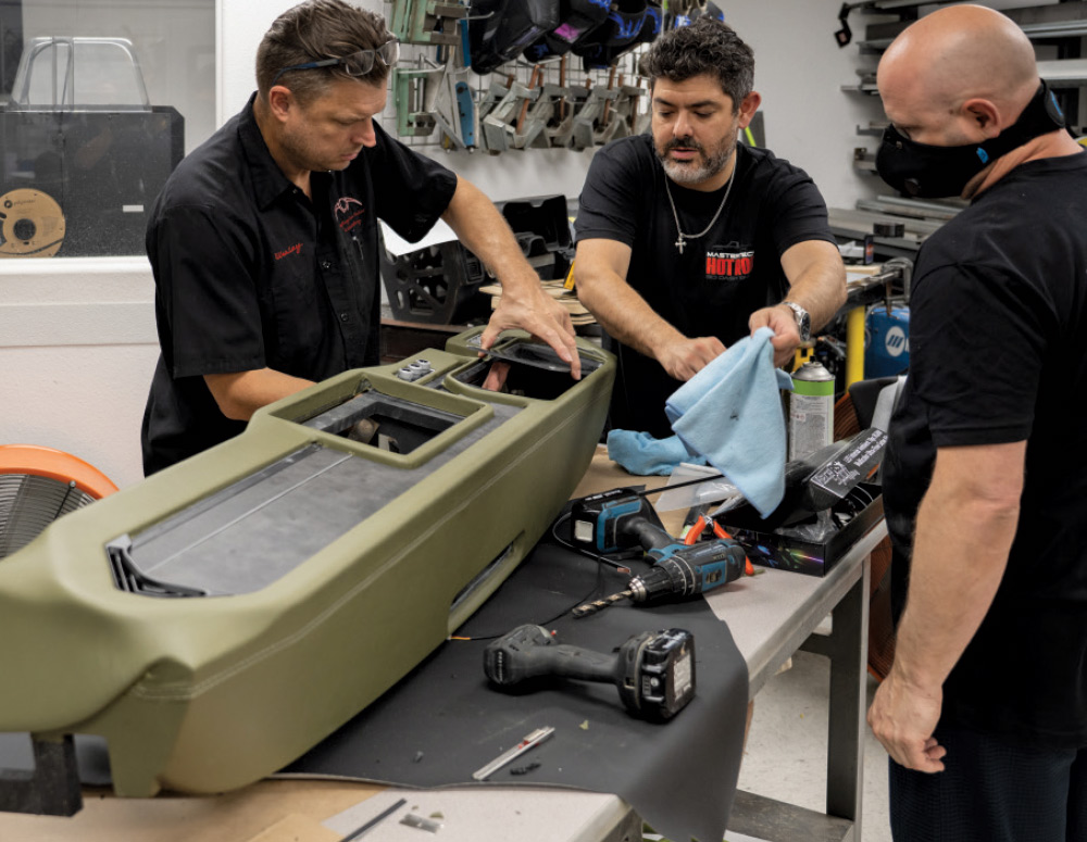 Three men are working on a large, custom vehicle dashboard wrapped in olive green vinyl or leather on a workbench. The man on the left is fitting a black panel into the dash. The man in the center is holding a light blue cloth and looking down at the work. The third man who is wearing a face mask around his neck, stands to the right. Power drills and other tools are on the table in the foreground.