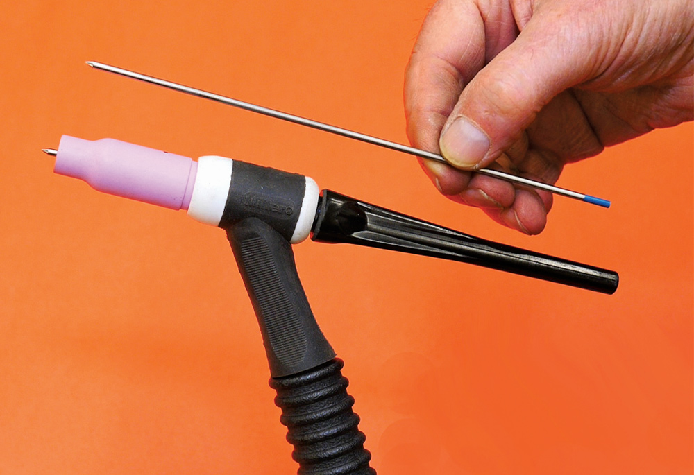 TIG torch holding blue-tip tungsten, pink cup, operator positioning electrode, orange background
