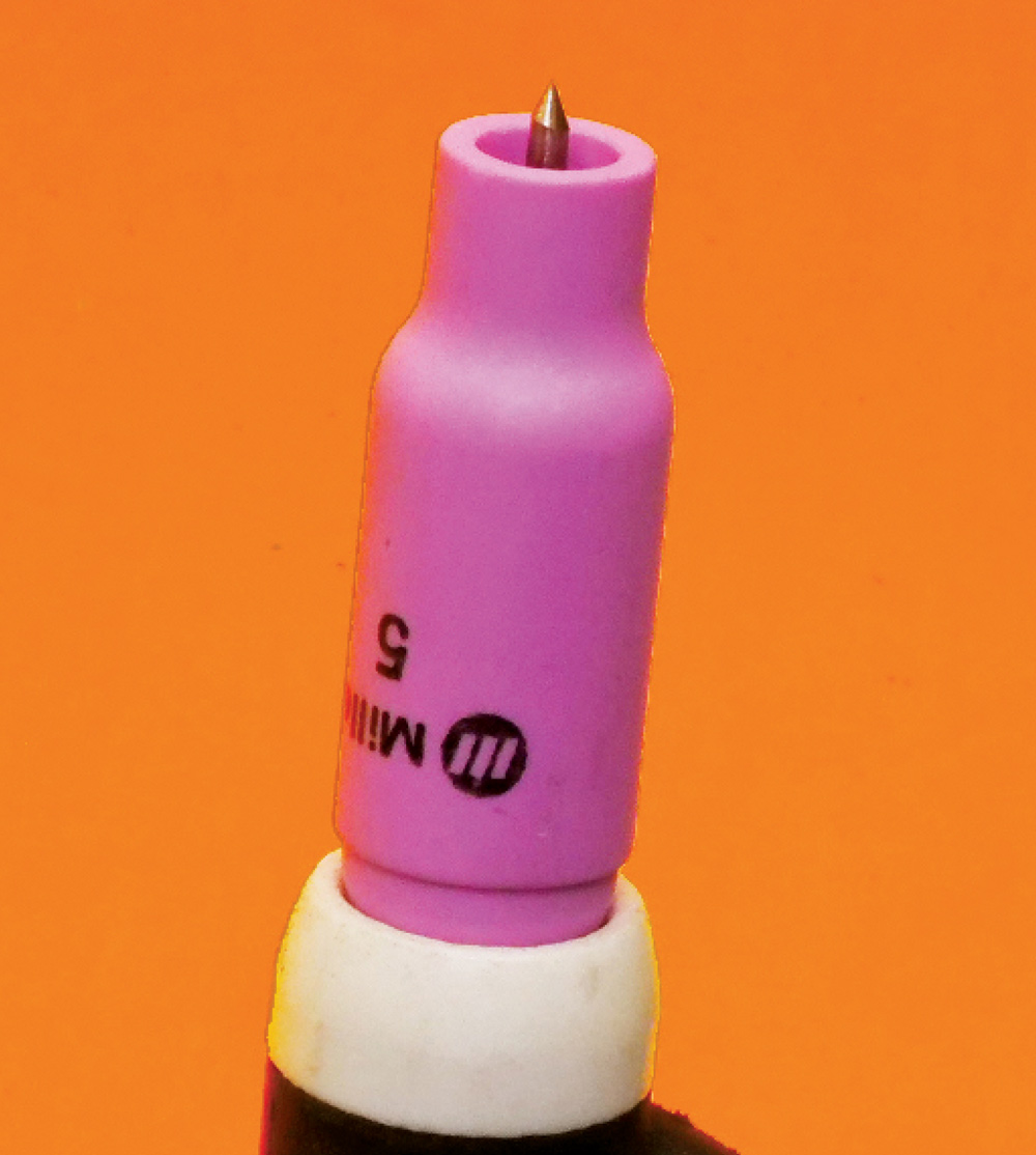 TIG torch close-up, pink MILLER #5 cup, tungsten tip, white band below cup, orange background