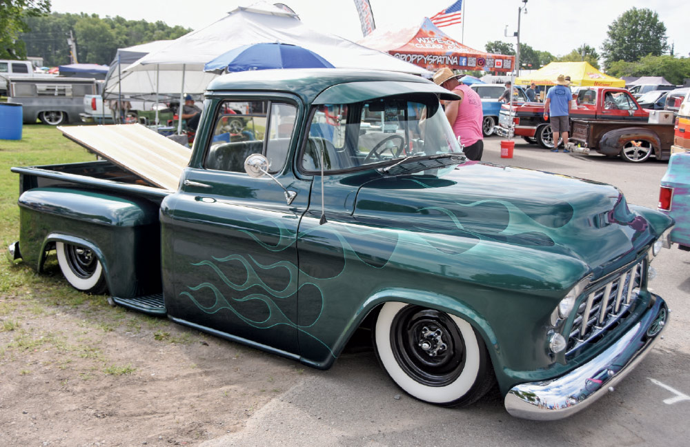A low-riding, custom dark green classic pickup truck with subtle ghost flames painted on the hood, white-wall tires, and a wooden truck bed at a car show.