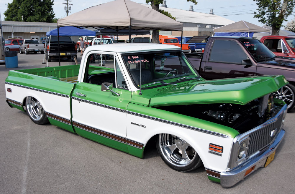 A custom, lowered green and white Chevrolet C10 pickup truck with its hood open and chrome rims, displayed at an outdoor car show.