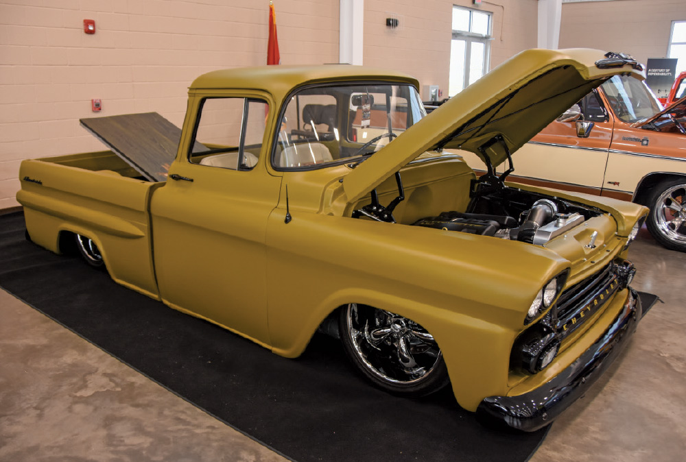 A classic, lowered matte mustard-yellow Chevrolet pickup truck on chrome rims, displayed indoors with the hood and bed cover open.