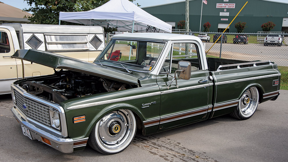 A custom, low-riding green and white Chevrolet C10 pickup truck with chrome wheels and the hood open, displayed outdoors at a venue.