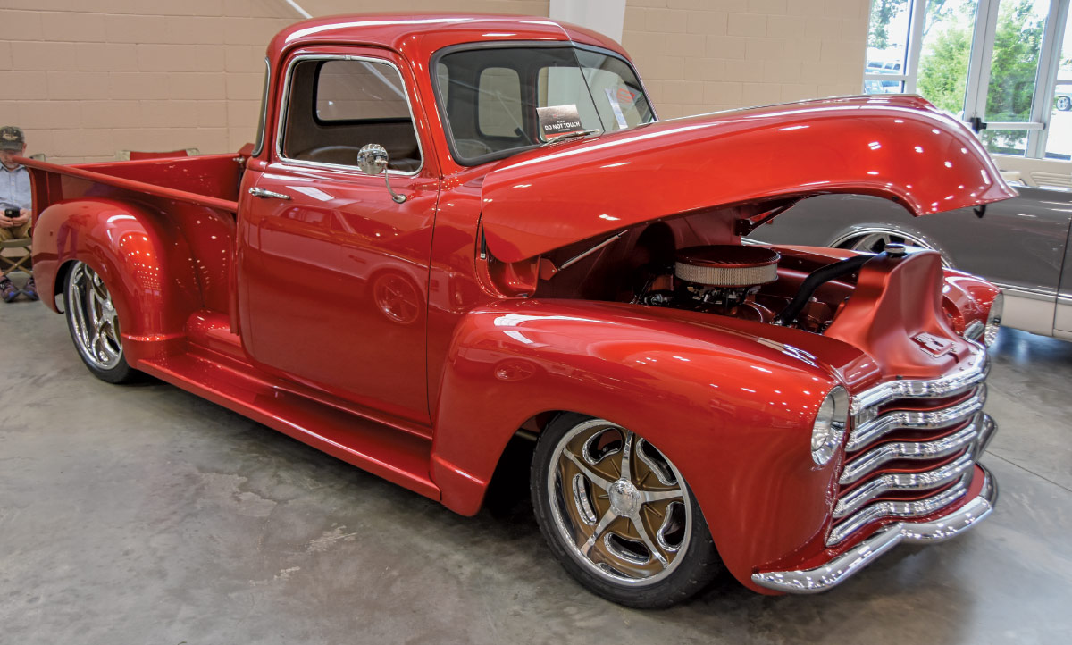 A shiny, customized red classic Chevrolet pickup truck displayed indoors with the hood open, revealing the engine, and chrome rims.